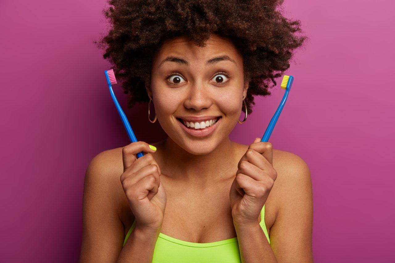 Dental hygiene concept. Glad surprised Afro American woman holds two tooth brushes, smiles broadly, brushes teeth in morning, has bare shoulders, clean dark skin isolated over lilac background.